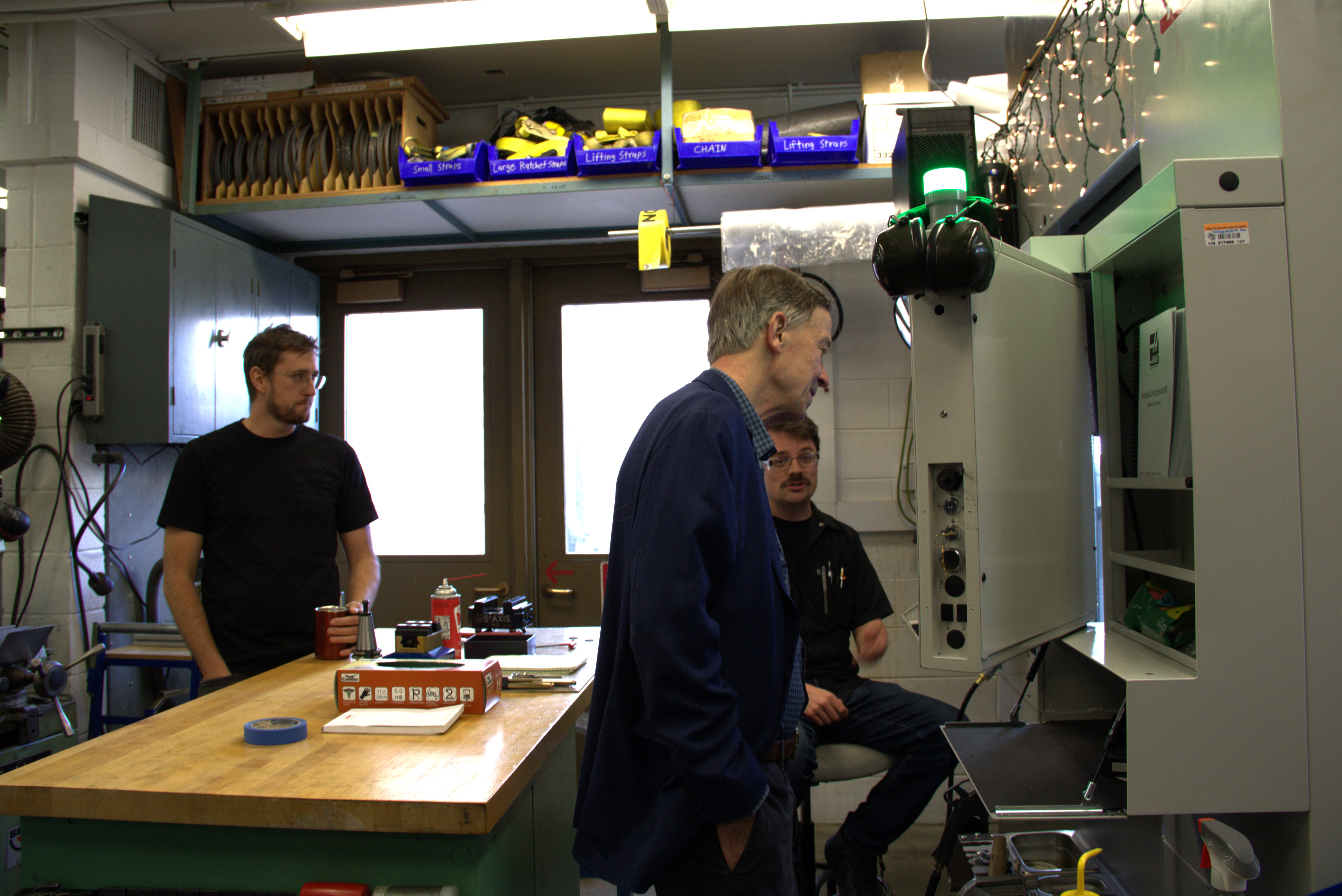 Senator Hickenlooper (center), examines JILA's new milling machine while instrument shop head Kyle Thatcher (left), and instrument shop apprentice Adam Elzey (right), watch