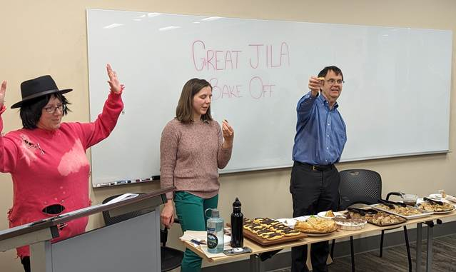 (left to right): Beth Kroger, aka "Noel Fielding," announces a new bake as the judges, graduate student Iona Binne, and JILA and NIST Fellow Eric Cornell, listen